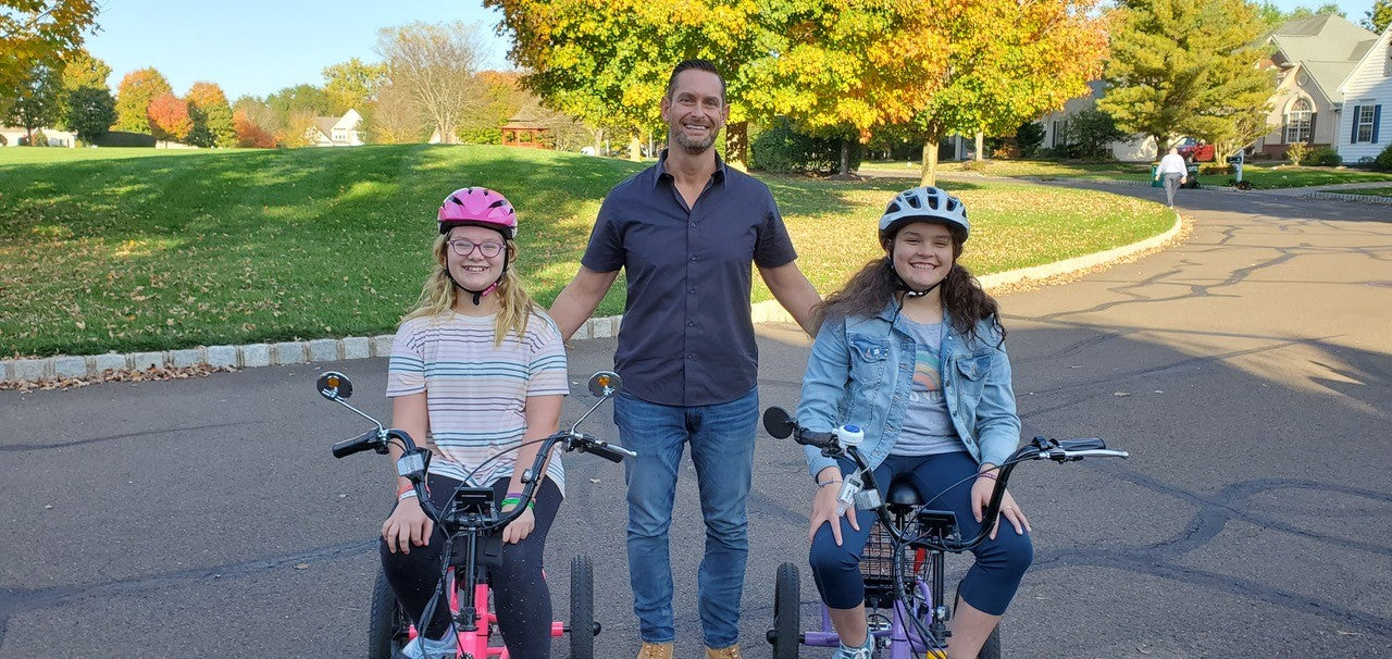 Two girls are sitting on a bicycle, and a man is standing by.